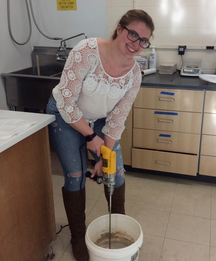 white woman with glasses and lace shirt mixes a bucket of clay with a drill